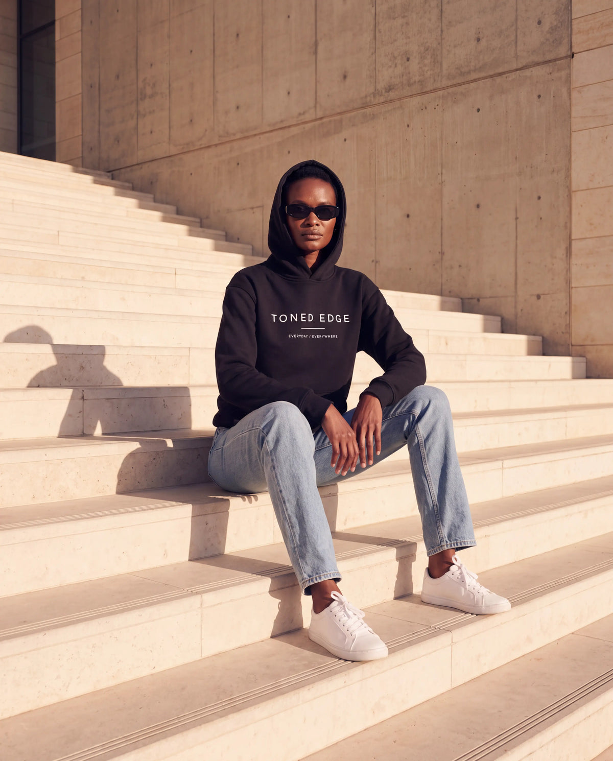 The Everyday Hoodie in Black, seated on concrete steps